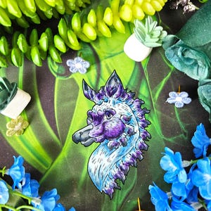 May include: A decorative pin featuring a stylized creature head with a pointed snout, large ears, and a mane, colored in shades of white, light blue, and deep purple. The creature has a dark beak-like nose and expressive eyes with black outlines.