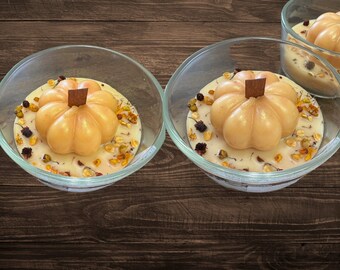 May include: Three glass candle jars with a pumpkin-shaped candle inside each. The candles are a light orange color and have a wooden stem. The jars are filled with dried flowers and other natural elements.