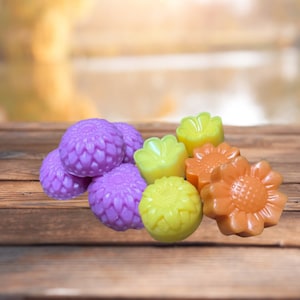 May include: A collection of colorful, flower-shaped soaps. The soaps are in shades of purple, yellow, and orange. The soaps are arranged on a wooden surface with a blurred background of a lake and trees.