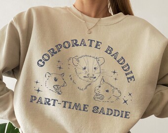 Könnte beinhalten: Ein beiger Pullover mit einem grafischen Druck von drei Opossums und dem Text "Corporate Baddie Part-Time Saddie".