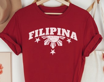 May include: Red t-shirt with a white graphic of the Philippine sun and stars with the word "FILIPINA" in a white vintage collegiate font.
