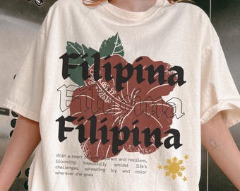 May include: Ivory-colored t-shirt with the word "Filipina" in a bold, stylized font, along with a floral graphic. The shirt also features text about blooming beautifully and spreading joy.