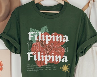 May include: Olive green t-shirt with a red hibiscus flower graphic and the word "Filipina" repeated three times in a vintage font. The text below the graphic reads "With a heart of gold vibrant and resilient, blooming beautifully amidst life's challenges, spreading joy and color wherever she goes."