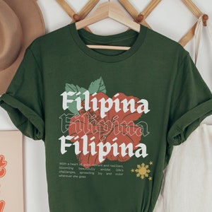 May include: Olive green t-shirt featuring a red hibiscus flower graphic and the word "Filipina" repeated three times in a vintage font. The text below the graphic reads "With a heart of gold vibrant and resilient, blooming beautifully amidst life's challenges, spreading joy and colour wherever she goes."