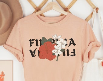 May include: A soft pink t-shirt with a floral design and the text "FLORIA" printed in black.