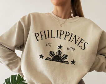 May include: A beige sweatshirt with a black graphic print of the words "PHILIPPINES EST 1899" and a stylized sun with stars. The graphic is reminiscent of the Philippine flag.