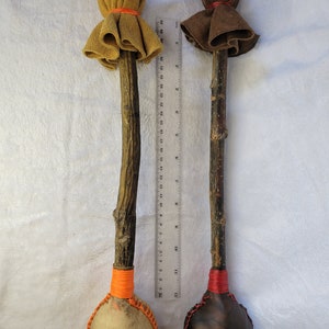 May include: Two handmade rattles with wooden handles and leather coverings. The rattles are tied with orange and red string. The rattles are approximately 30 cm long.