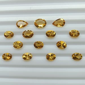 May include: A collection of polished, golden-yellow gemstones in various shapes, including oval and pear cuts. The gems are arranged on a white, ridged surface, showcasing their clarity and brilliance. The gemstones are likely citrine.