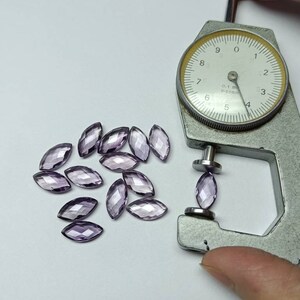 Peut inclure: Plusieurs pierres précieuses taillées en marquise, de couleur violet clair, sont disposées sur une surface blanche. Un pied à coulisse à cadran mesure l'épaisseur d'une pierre. Le pied à coulisse est argenté.