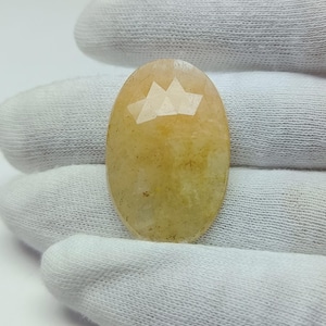 Peut inclure: Une pierre précieuse ovale et facettée, de couleur jaune-orangé clair. La gemme a une surface polie avec un motif géométrique sur le dessus. La pierre est tenue par un tissu blanc.