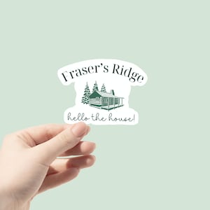 May include: A green and white sticker with the text "Fraser's Ridge" and "hello the house!"  A drawing of a cabin with trees is in the center of the sticker.