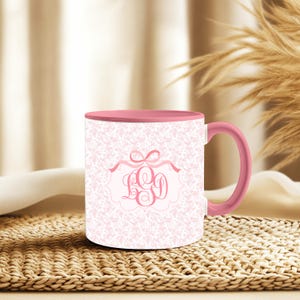May include: A pink and white ceramic mug with a floral pattern and a pink handle. The mug features a decorative label with the letters LGD in a script font, framed by a ribbon and bow design. The interior of the mug is also pink.