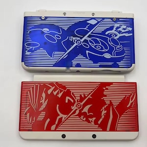 May include: Two Nintendo 3DS consoles. One is blue with a white outline of a creature and the other is red with a white outline of a creature. Both consoles have a white base and a horizontal striped design.