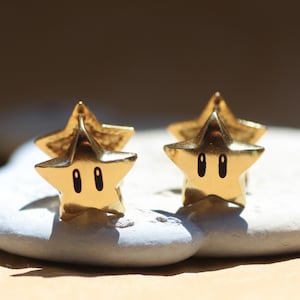 May include: Gold-colored star-shaped earrings with black oval eyes. The earrings are on a white stone, with a blurred background. The earrings are a stylized representation of a star.