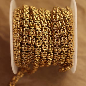 Puede incluir: Un carrete de cadena dorada. La cadena está hecha de pequeños eslabones rectangulares que están estrechamente conectados. La cadena está enrollada alrededor de una bobina de plástico blanco. El fondo es de color marrón claro.