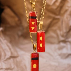 May include: Three gold-toned necklaces featuring rectangular pendants with a glossy red finish. Each pendant displays distinct gold-colored geometric symbols, including two downward-pointing triangles, two diamonds, and a single circle, with black bands.