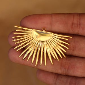 以下が含まれることがあります： 半円形のトップと放射状のスパイクを備えた、金色のサンバーストペンダント。ペンダントの上部には、吊り下げ用の小さなループが付いています。 デザインは対称的で詳細で、研磨された仕上げです。