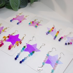 May include: Assortment of colorful beaded dangle earrings displayed on white cards with a star design. Each earring features a variety of crystal beads in different colors and shapes.