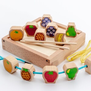 May include: A wooden toy set with colourful fruit-shaped beads and a wooden box with a lid. The beads can be strung on a blue cord or a yellow cord. The box has two wooden sticks for playing.