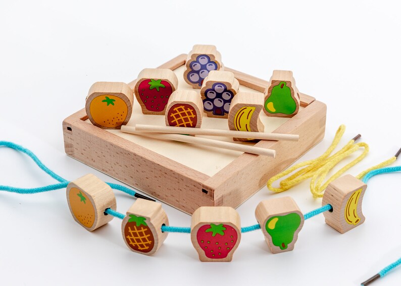 May include: A wooden toy set with colourful fruit-shaped beads and a wooden box with a lid. The beads can be strung on a blue cord or a yellow cord. The box has two wooden sticks for playing.