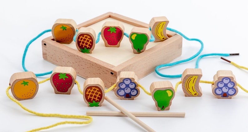 May include: A wooden toy with colourful fruit-shaped beads that can be strung on a string. The beads include an orange, pineapple, strawberry, pear, banana, and grapes. The toy comes with a wooden box and two wooden sticks.