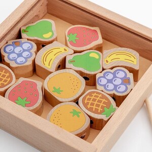 May include: A wooden box filled with colourful wooden fruit shapes. The fruit shapes have holes in them and are used for threading string. There are two wooden needles and a length of string in the image.
