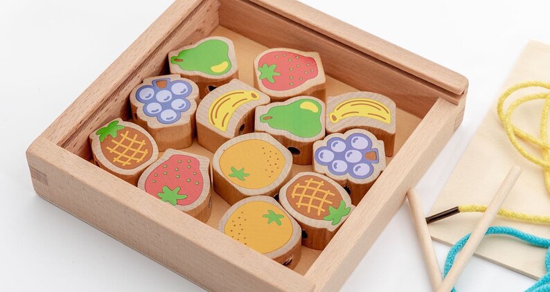 May include: A wooden box filled with colourful wooden fruit shapes. The fruit shapes have holes in them and are used for threading string. There are two wooden needles and a length of string in the image.