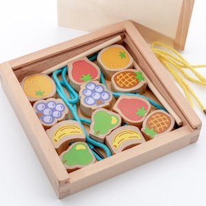 May include: A wooden box with a lid contains a set of colourful wooden fruit shapes with string and a wooden needle. The fruit shapes include strawberries, pineapples, bananas, pears, grapes, and oranges.