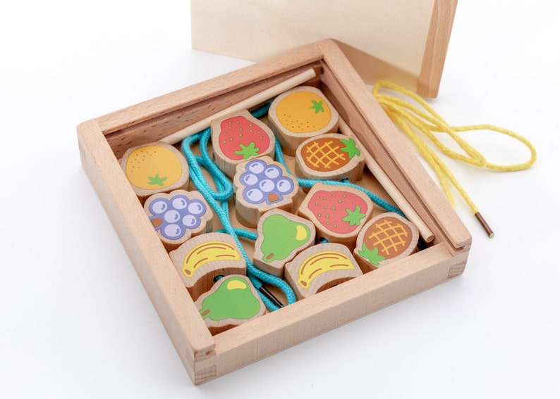 May include: A wooden box with a lid contains a set of colourful wooden fruit shapes with string and a wooden needle. The fruit shapes include strawberries, pineapples, bananas, pears, grapes, and oranges.