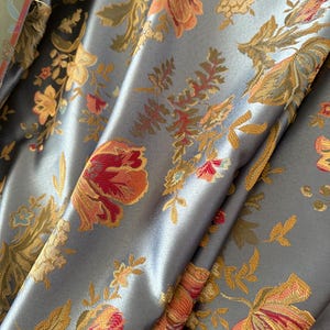 Può includere: Primo piano di un tessuto lussuoso con motivo floreale. Il tessuto è di un morbido colore blu argentato, con una lucentezza. Il design floreale presenta fiori e foglie arancioni, rosse e dorate, creando un aspetto ricco e strutturato.