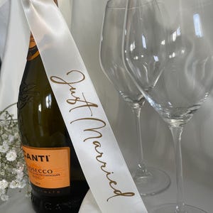 以下が含まれることがあります： 「Just Married」と金色の文字で書かれた白いリボンが付いたプロセッコのボトル。背景には2つの透明なワイングラスがあります。ボトルにはオレンジ色のラベルが付いており、白いリボンで結ばれています。