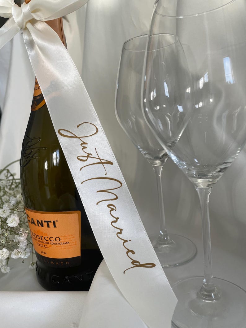 以下が含まれることがあります： 「Just Married」と金色の文字で書かれた白いリボンが付いたプロセッコのボトル。背景には2つの透明なワイングラスがあります。ボトルにはオレンジ色のラベルが付いており、白いリボンで結ばれています。