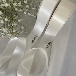 以下が含まれることがあります： 白とアイボリーのサテンリボンのロールで、リボンには「WHITE」と「IVORY」の文字がプリントされています。リボンは白い花の小枝を背景に配置されています。リボンは光沢のある仕上がりです。