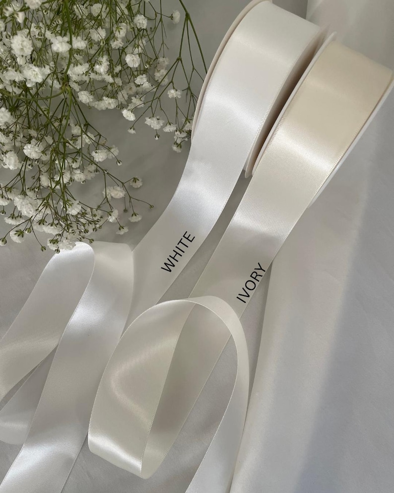以下が含まれることがあります： 白とアイボリーのサテンリボンのロールで、リボンには「WHITE」と「IVORY」の文字がプリントされています。リボンは白い花の小枝を背景に配置されています。リボンは光沢のある仕上がりです。