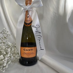 以下が含まれることがあります： 白いサテンのリボンで飾られた、Canti Proseccoのボトル。リボンには「Just Married」の文字が入ったタグが付いています。ボトルにはオレンジ色のラベルが付いています。