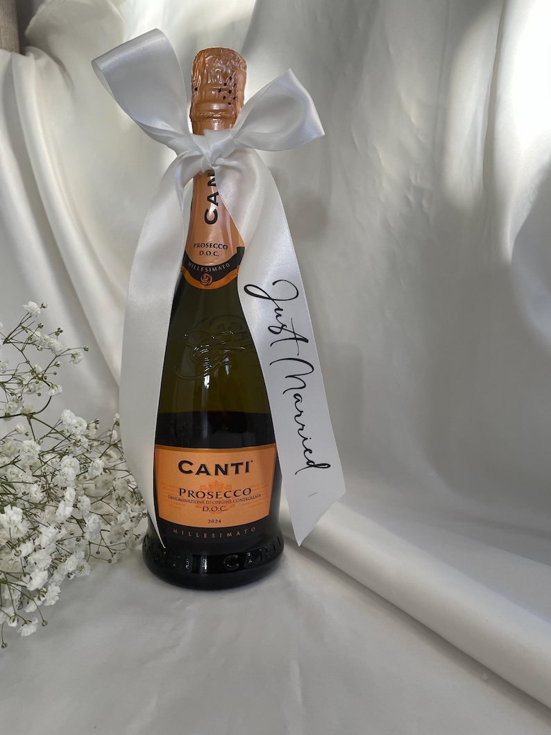 以下が含まれることがあります： 白いサテンのリボンで飾られた、Canti Proseccoのボトル。リボンには「Just Married」の文字が入ったタグが付いています。ボトルにはオレンジ色のラベルが付いています。