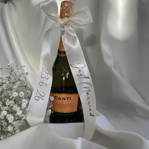 以下が含まれることがあります： 白いサテンのリボンが首に巻かれた、Canti Proseccoのボトル。リボンには「Just Married」と日付「23.6.26」が金色の文字で印刷されています。ボトルは白い背景に置かれています。