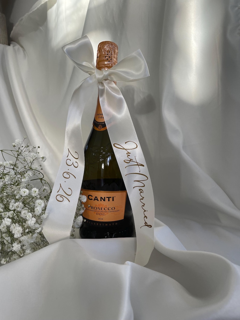以下が含まれることがあります： 白いサテンのリボンが首に巻かれた、Canti Proseccoのボトル。リボンには「Just Married」と日付「23.6.26」が金色の文字で印刷されています。ボトルは白い背景に置かれています。