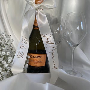 以下が含まれることがあります： 白いサテンのリボンが巻かれたCanti Proseccoワインのボトル。リボンには「Just Married」と日付「23.6.26」と書かれています。ボトルの隣にはワイングラスが2つあります。