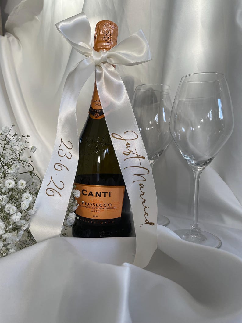 以下が含まれることがあります： 白いサテンのリボンが巻かれたCanti Proseccoワインのボトル。リボンには「Just Married」と日付「23.6.26」と書かれています。ボトルの隣にはワイングラスが2つあります。