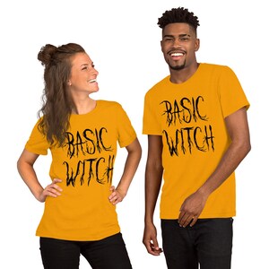以下が含まれることがあります： 2人の人が、黒い文字で「BASIC WITCH」とプリントされた黄色のTシャツを着ています。テキストはゴシック体でスタイリッシュです。