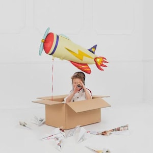 May include: A child in a cardboard box with paper airplanes scattered around. A large, colorful airplane balloon is suspended above. The balloon is yellow, red, and blue with a yellow lightning bolt design. The child is wearing goggles and looking through a tube.