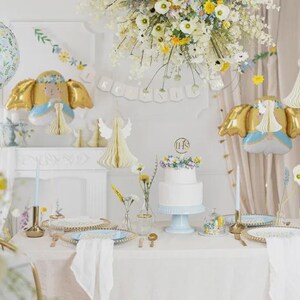 May include: A table setting featuring a white two-tier cake with a gold IHS topper, blue plates, gold cutlery, and white napkins. Decorations include angel-shaped foil balloons, white paper honeycomb angels, and a floral balloon with text.