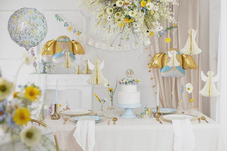 May include: A table setting featuring a white two-tier cake with a gold IHS topper, blue plates, gold cutlery, and white napkins. Decorations include angel-shaped foil balloons, white paper honeycomb angels, and a floral balloon with text.