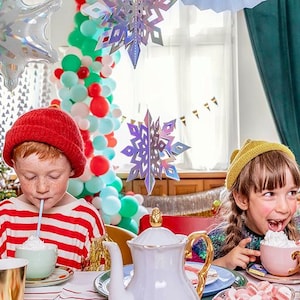 Puede incluir: Dos ni&ntilde;os en una mesa con una tetera blanca, tazas pastel con nata montada y pajitas. Un ni&ntilde;o lleva un gorro rojo y una camiseta a rayas, el otro un gorro amarillo. El entorno festivo incluye un &aacute;rbol de Navidad y copos de nieve colgantes.