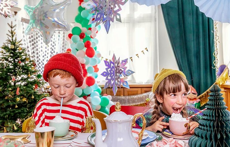 Puede incluir: Dos ni&ntilde;os en una mesa con una tetera blanca, tazas pastel con nata montada y pajitas. Un ni&ntilde;o lleva un gorro rojo y una camiseta a rayas, el otro un gorro amarillo. El entorno festivo incluye un &aacute;rbol de Navidad y copos de nieve colgantes.