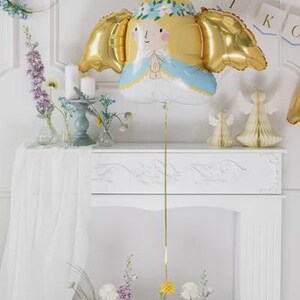 May include: A foil balloon shaped like an angel with blonde hair, a blue and white robe, and metallic gold wings. The angel has a floral crown and hands pressed together in prayer. A thin gold ribbon hangs from the bottom of the balloon.