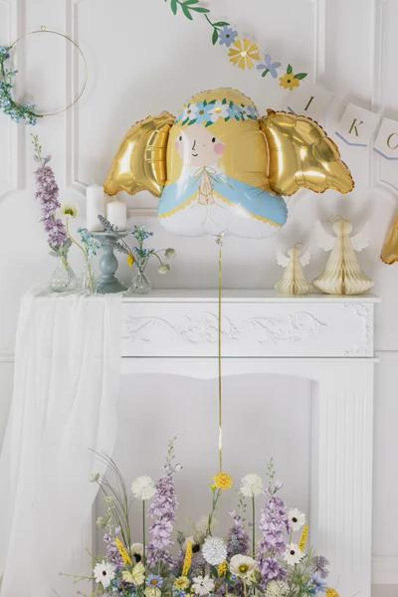 May include: A foil balloon shaped like an angel with blonde hair, a blue and white robe, and metallic gold wings. The angel has a floral crown and hands pressed together in prayer. A thin gold ribbon hangs from the bottom of the balloon.