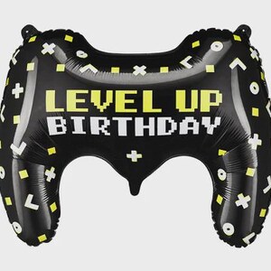 May include: A black foil balloon shaped like a video game controller, featuring the text "LEVEL UP BIRTHDAY" in yellow and white pixelated font. The surface is decorated with a pattern of small yellow squares, white plus signs, and circles.