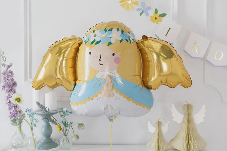 May include: A foil balloon shaped like a stylised angel with blonde hair, a blue and white robe, and large gold wings. The figure has a floral crown, pink cheeks, and hands pressed together in a prayer position, attached to a thin gold string.